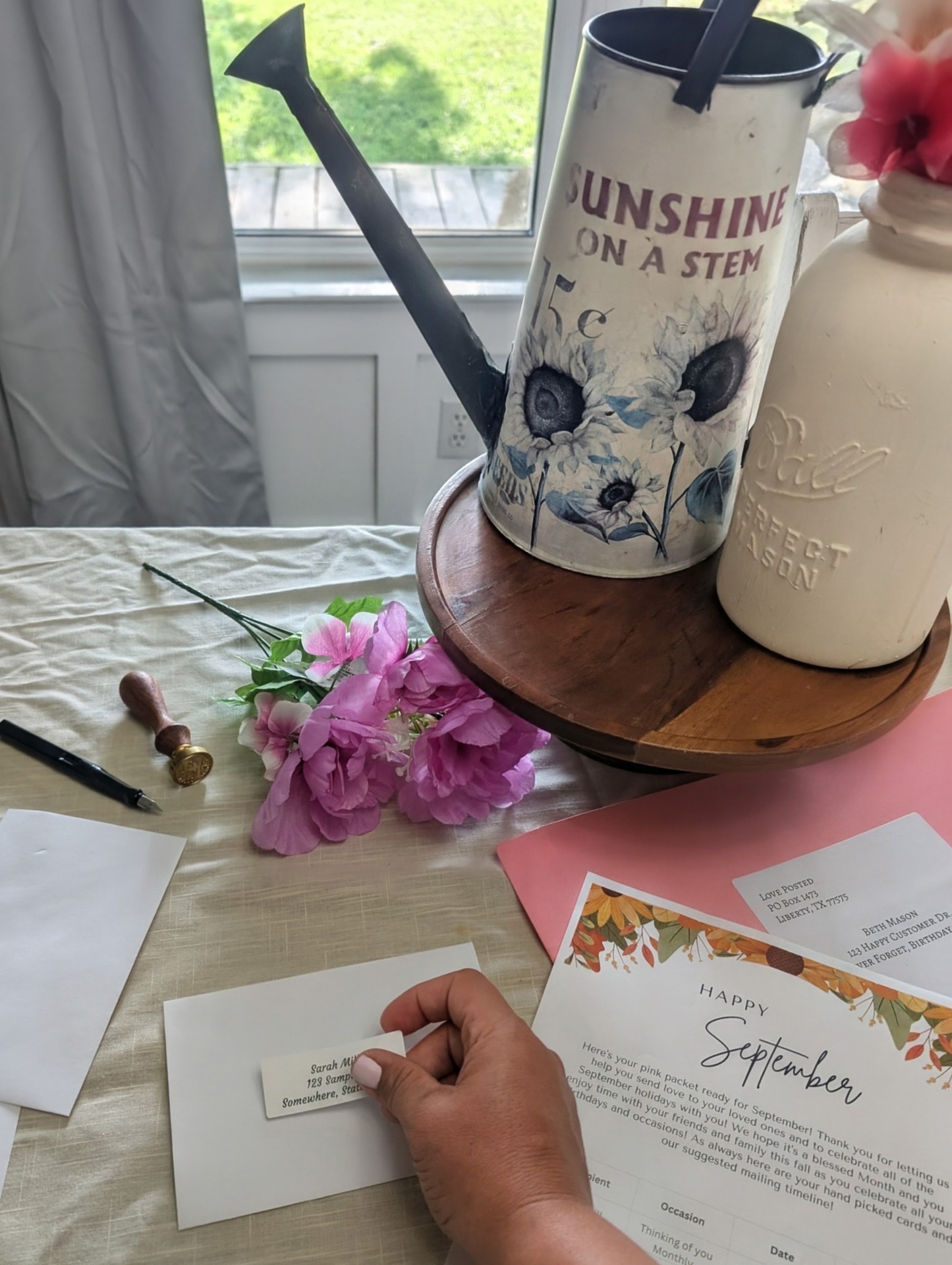 Madisson's hand placing a printed address label onto a white card envelope, beside the pink packet and the monthly fridge sheet