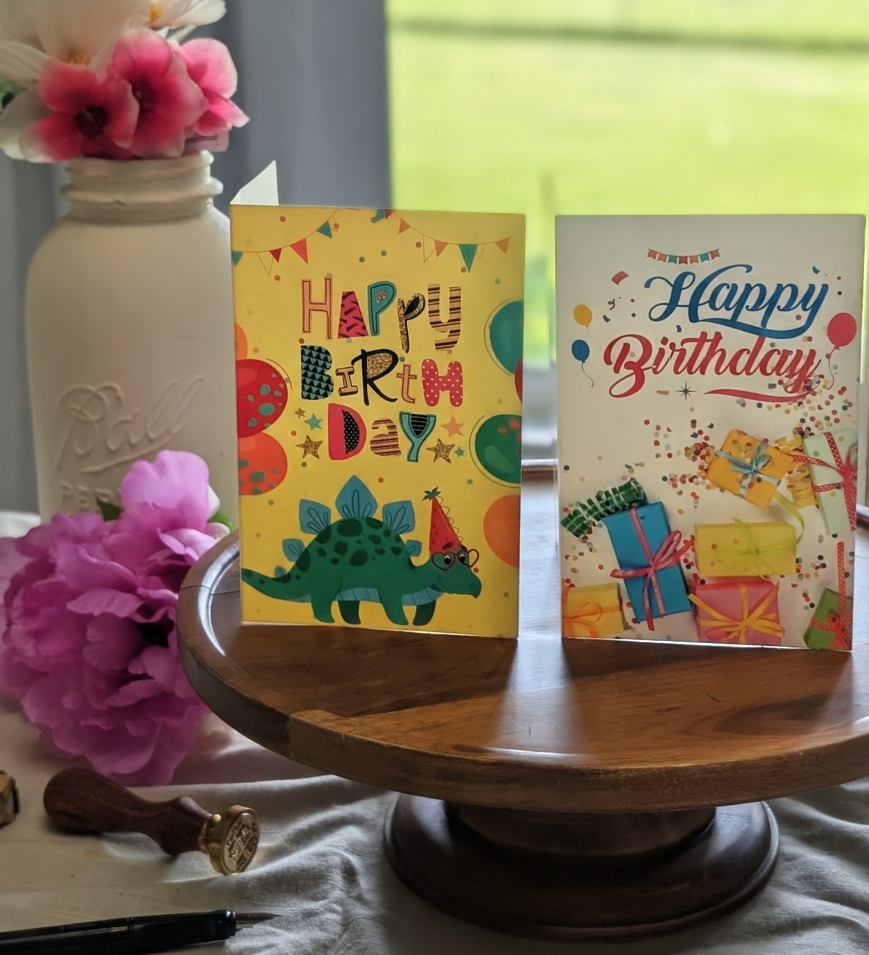 Two real birthday cards standing side by side — a bright yellow kid card with a party-hat dinosaur, and a white adult card with hand-lettered script and confetti