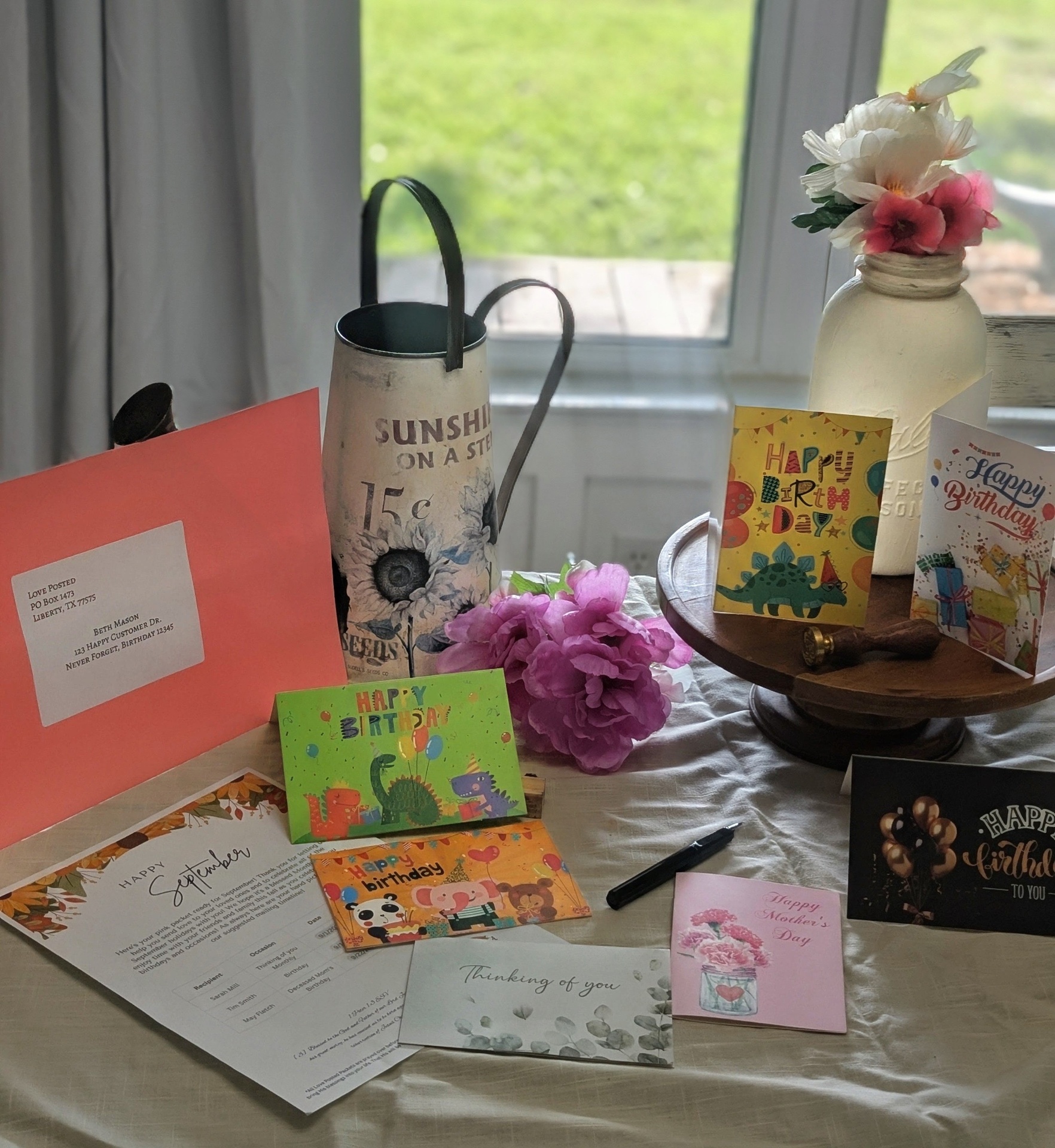 A full Love Posted packet spread across a table — birthday cards, a Mother's Day card, a thinking-of-you card, the pink packet envelope, and the monthly fridge sheet