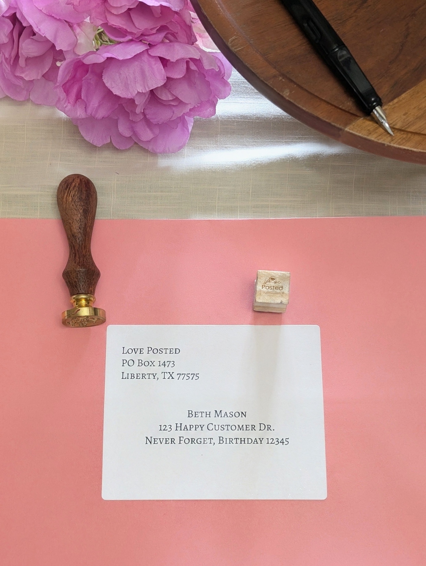 The coral pink Love Posted packet with its printed mailing label, a wax seal stamp, and the Love Posted rubber stamp on a linen table